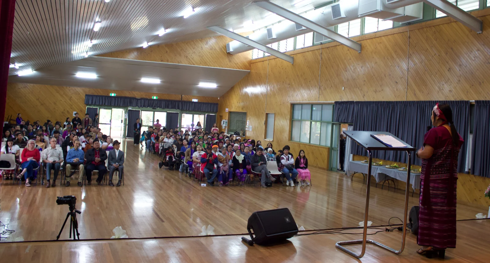 A woman in traditional Karen dress speaking at a lectern to a hall full of people at the wrist-tying ceremony.
